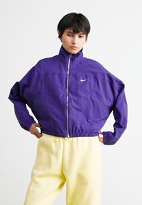 Žmogus, vilkintis violetinę Nike zip-up striukę ir šviesiai geltonas Nike sportines kelnes, stovi su rankomis ant klubų prieš paprastą baltą foną.