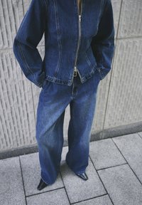 Osoba u tamnoplavoj traper jakni i širokim trapericama, s rukama u džepovima, stoji na pločniku s pločicama blizu teksturiranog zida.