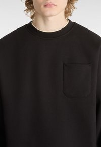 Sweat-shirt noir avec un col rond, un tissu texturé et une petite poche poitrine à bord arrondi.