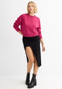 Suéter de punto fucsia con cuello redondo, combinado con una falda envolvente negra que presenta una abertura alta y una tela texturizada. Botines negros de tacón.