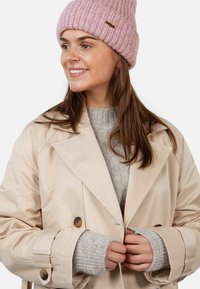 Young woman smiling, wearing a pink knit hat, beige trench coat, and gray knitted sweater, adjusting her coat buttons.