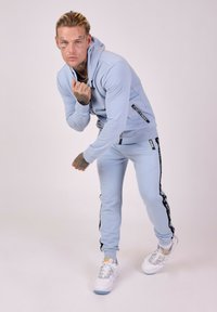 Lichtblauwe trainingspak met een hoodie, voorzien van zwarte zijstrepen versierd met tekst. Compleet met witte sportschoenen.