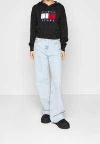 Person wearing black cable-knit Tommy Jeans hoodie, light blue wide-leg jeans, and black platform shoes standing against a plain background.
