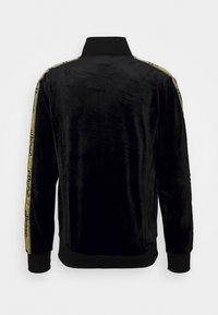 Svart sammet bomberjacka med långa ärmar, ribbad krage och muddar. Har gulddetaljer längs sidorna med logotext. Slät textur.