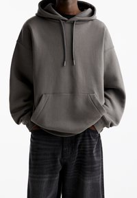 Person som bär en mörkgrå huva med framficka och svarta jeans, stående med händerna i fickorna mot en vit bakgrund.