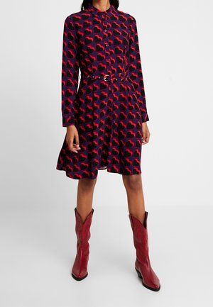 Mujer vestida con un vestido azul marino con patrones de corazones rojos, con cintura ceñida con cinturón y botas de vaquero rojas hasta mitad de la pantorrilla, de pie contra un fondo liso.