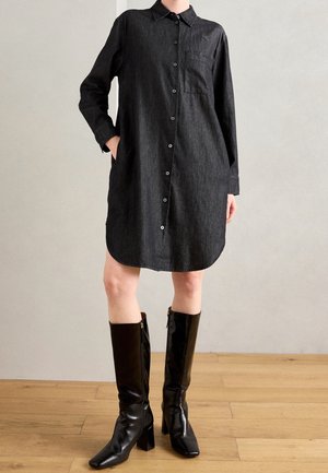 Personne portant une robe chemise noire boutonnée avec une main dans la poche et des bottes noires à talons montant jusqu'aux genoux, debout sur un sol en bois.
