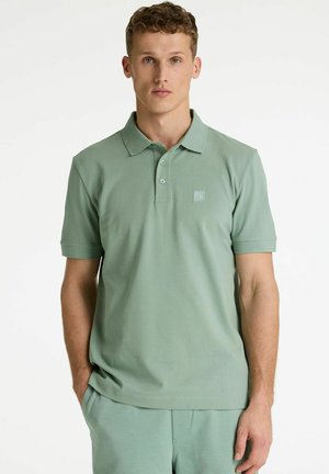 Młody mężczyzna ubrany w jasnozieloną koszulkę polo z krótkim rękawem i małym logo na lewej piersi, stojący na tle jednolitego, białego tła.