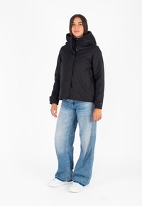 Zwarte gewatteerde jas met een capuchon, voorzien van een knoopsluiting aan de voorkant en twee zijzakken, gedragen over lichtblauwe wijde jeans en witte sneakers.