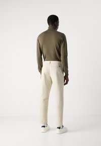 Olive zaļš augstais kakls, krēmkrāsas bikses, slim fit, gluda auduma un baltas sporta apavi ar melniem akcentiem. Nav redzama zīmola identifikācija.