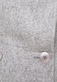 Grijs, textielachtig materiaal met een subtiel patroon, voorzien van een metalen knoop aan de linkerkant. Het oppervlak lijkt zacht en lichtjes donsig.