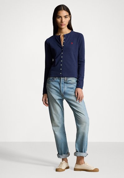 Chemise boutonnée à manches longues de couleur navy avec un col rond et un petit logo, associée à un jean clair à coupe décontractée et des baskets beiges.