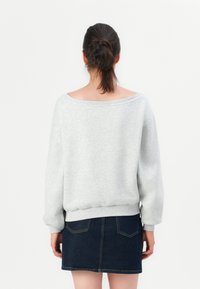 Donna con capelli scuri raccolti, che indossa un maglione grigio chiaro e una gonna di jeans blu scuro, vista da dietro su uno sfondo semplice.