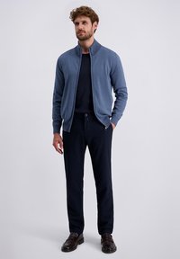 Mann steht da, trägt einen blauen Windbreaker über einem schwarzen Shirt, schwarze Hosen und dunkelbraune Schuhe vor einem schlichten hellen Hintergrund.