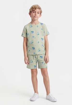 Ung dreng står iført lysegrøn matchende kortærmet skjorte og shorts med tropiske øer og dyreprints, hvide sneakers.