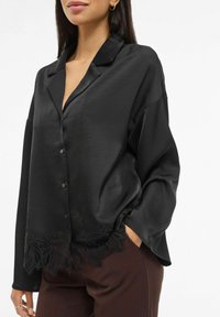 Svart satengbluse med krage, lange ermer, knapper foran og blondedetaljer i kanten. Jevn tekstur, ledig passform.