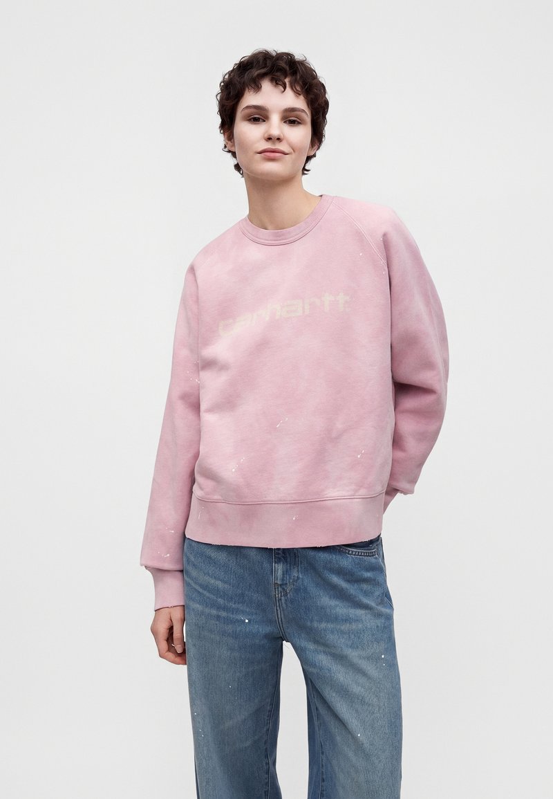 Junge Person mit kurzen dunklen Haaren, die einen hellrosa Sweatshirt und blaue Jeans trägt, steht mit einer Hand in der Tasche vor einem schlichten Hintergrund.
