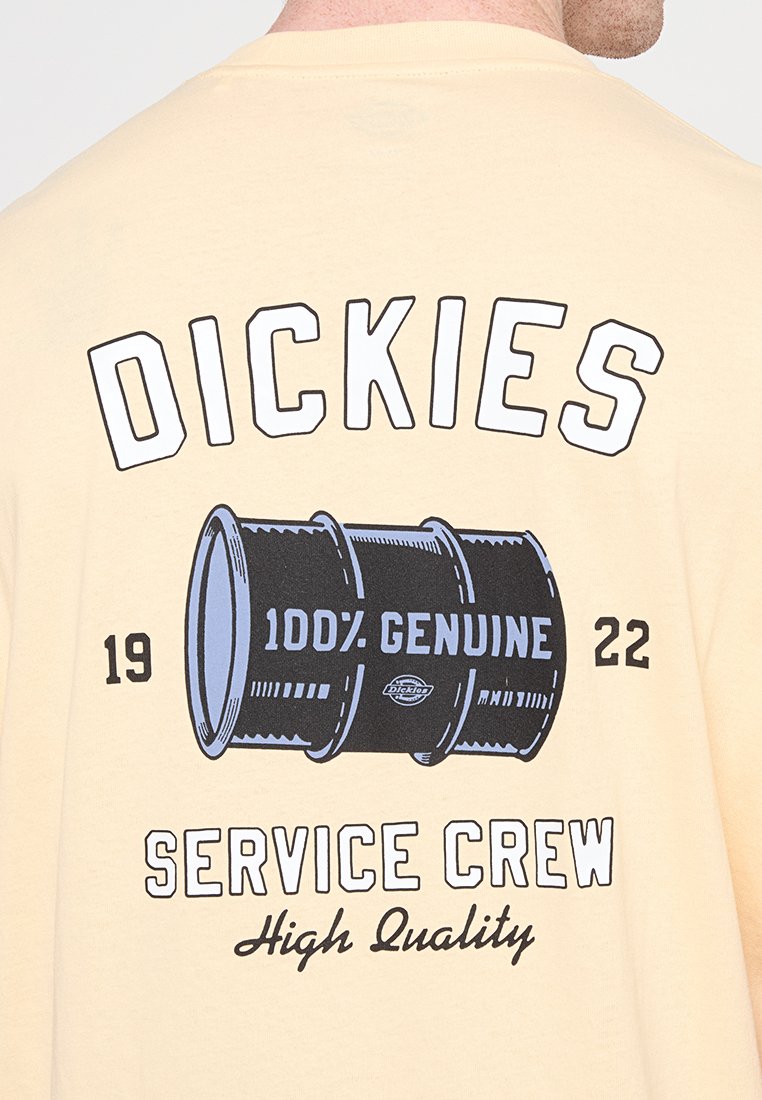 Persona che indossa una camicia beige con grafica di una botte nera e la scritta "Dickies 100% Genuine 1922 Service Crew High Quality" sul retro.