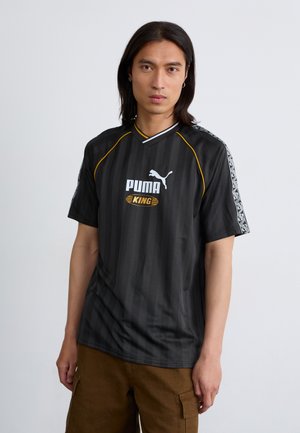A young man stands confidently, wearing a dark striped Puma King jersey with yellow accents and brown cargo pants, against a light background.