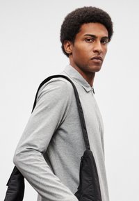 Γκρι μπλούζα πόλο με μακριά μανίκια και κολάρο με φερμουάρ, φορεμένη με μαύρη τσάντα χιαστί με υφή καπιτονέ και λεπτομέρεια φερμουάρ.