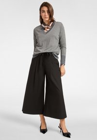 Grijs gebreide trui met V-hals en lange mouwen, gedragen over een meerkleurig overhemd met kraag, gecombineerd met zwarte broek met wijde pijpen en puntige schoenen.