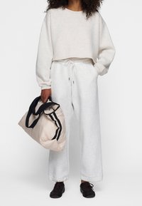 Persona in un maglione corto beige chiaro e pantaloni della tuta bianchi con cordoncino tiene una borsa a mano beige con manici neri, indossando scarpe nere.