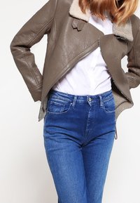 Veste en cuir marron avec col en shearling, fermeture éclair à l'avant, superposée sur une chemise blanche, associée à un jean bleu. Gros plan sur le haut du corps.