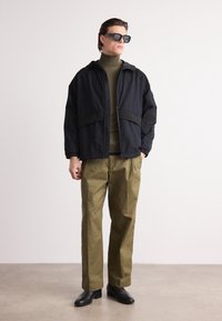Sort jakke med lynlås og kontrasterende paneler, olivengrøn turtleneck, khakifarvede bukser og sorte sko; stilede solbriller fuldender looket.