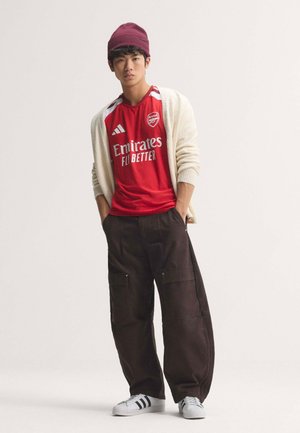 Ung mand iført en rød Arsenal fodboldtrøje, beige cardigan, vinrød hue, sorte cargobukser og hvide sneakers med sorte striber.