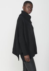 Črna pletena turtleneck pulover z ohlapnim krojem, rebrasto teksturo, visokim ovratnikom in stranskimi razporki. Združen z ozkimi črnimi hlačami.