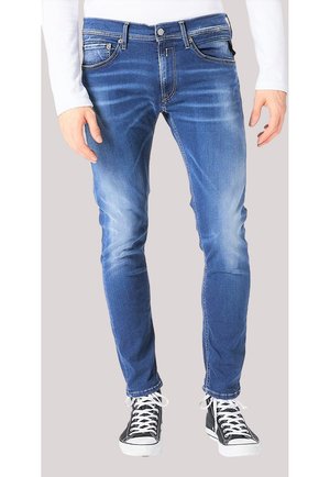 Schmal geschnittene blaue Jeans aus Denim mit ausgewaschenem Look, getragen von einer Person mit schwarz-weißen Sneakers und einem weißen Langarmshirt.