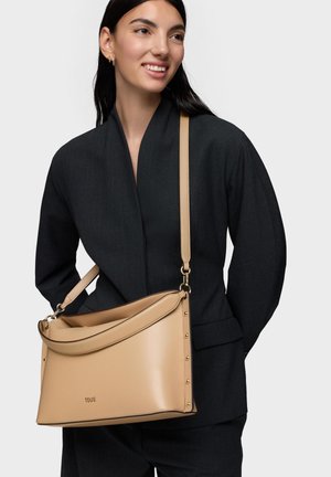 Mujer vistiendo un blazer y pantalones negros, llevando un bolso de hombro TOUS de cuero beige con detalles de tachuelas doradas.