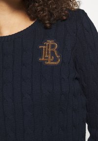 Marinblå kabelstickad tröja med ett strukturerat mönster och en brun broderad logotyp på vänster bröst. Rund halsringning och långa ärmar.