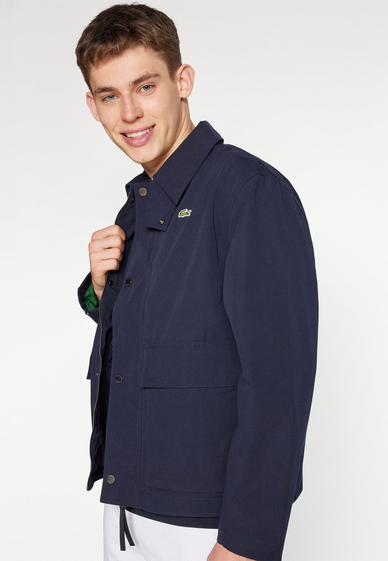 Giacca blu navy realizzata in tessuto resistente, con colletto classico, chiusura a bottoni, tasche frontali e un piccolo logo sul petto.