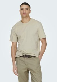 Beige Baumwoll-T-Shirt mit kurzen Ärmeln und Rundhalsausschnitt, kombiniert mit olivgrünen Hosen und einem braunen Gürtel, im schlichten Design.