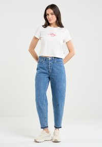 Weißes, lässig geschnittenes T-Shirt mit floralem roten Grafikdesign, kombiniert mit blauen, hochgeschnittenen Jeans und beigen Sneakers mit strukturierter Oberfläche.