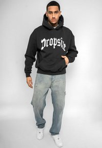 Svart hettegenser med hvit "Dropside" trykk, foran kengurulomme, kombinert med lyseblå slitte jeans og hvite joggesko.