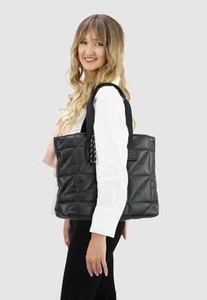 Lachende vrouw met lang blond haar draagt een grote zwarte gewatteerde tote tas over haar schouder, ze draagt een wit shirt en een zwarte broek.