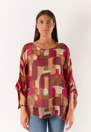 Donna che indossa una camicetta larga con motivo geometrico multicolore in tonalità di rosso, marrone e beige, in piedi davanti a uno sfondo bianco uniforme.