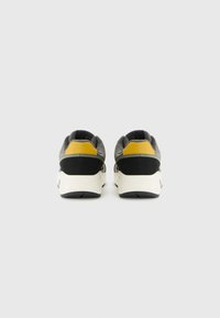 Une paire de baskets grises, noires et jaunes vue de l'arrière, avec des semelles blanches sur un fond blanc uni.
