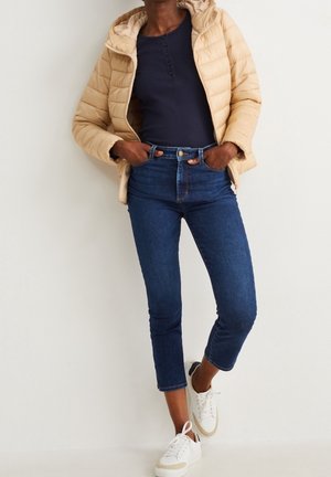 Personne portant une doudoune beige sur un t-shirt henley bleu marine, un jean court bleu foncé et des baskets blanches, debout les mains dans les poches.