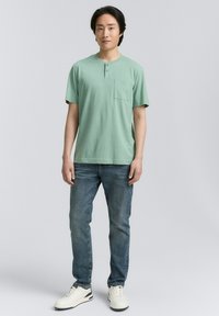 Tricou scurt de culoare verde pal, cu buzunar, purtat cu blugi albaștri și adidași albi. Tricoul are un gât rotund cu nasturi.