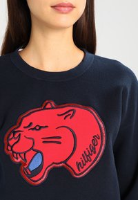 Sweat-shirt bleu marine avec un grand motif de panthère rouge brodé avec des accents bleus et le texte "hilfiger", tissu texturé et encolure côtelée.