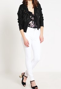 Černá umělá kožená bunda s květinovým topem, bílé skinny džíny a černé lodičky se zlatými akcenty. Modelka stojí s uvolněnými rukama.