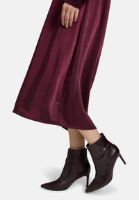 Femme portant une jupe midi bordeaux et des bottines à talons assorties avec des fermetures éclair latérales, se tenant devant un fond blanc.