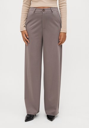 Kobieta ubrana w spodnie o wysokim stanie, szerokie nogawki w kolorze taupe, z zapięciem na guziki i zamek, w połączeniu z beżową bluzką z długim rękawem oraz czarnymi butami z zakrzywionymi noskami.