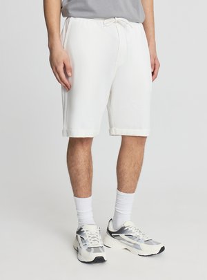 Mężczyzna ubrany w białe szorty z troczkiem, szarą koszulkę, białe skarpetki do kostki oraz szaro-białe buty sportowe, stojący na jednolitym tle.