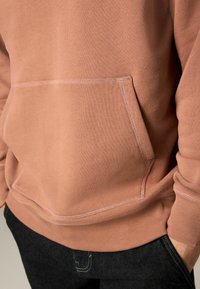 Light pink sweatshirt with a kangaroo pocket, ribbed cuffs, and hem. Soft fabric texture with visible stitching details. Worn with dark jeans.