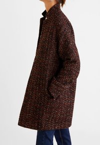 Abrigo texturizado y de gran tamaño en marrón oscuro con motas multicolores. Presenta un cuello, mangas largas y bolsillos laterales.