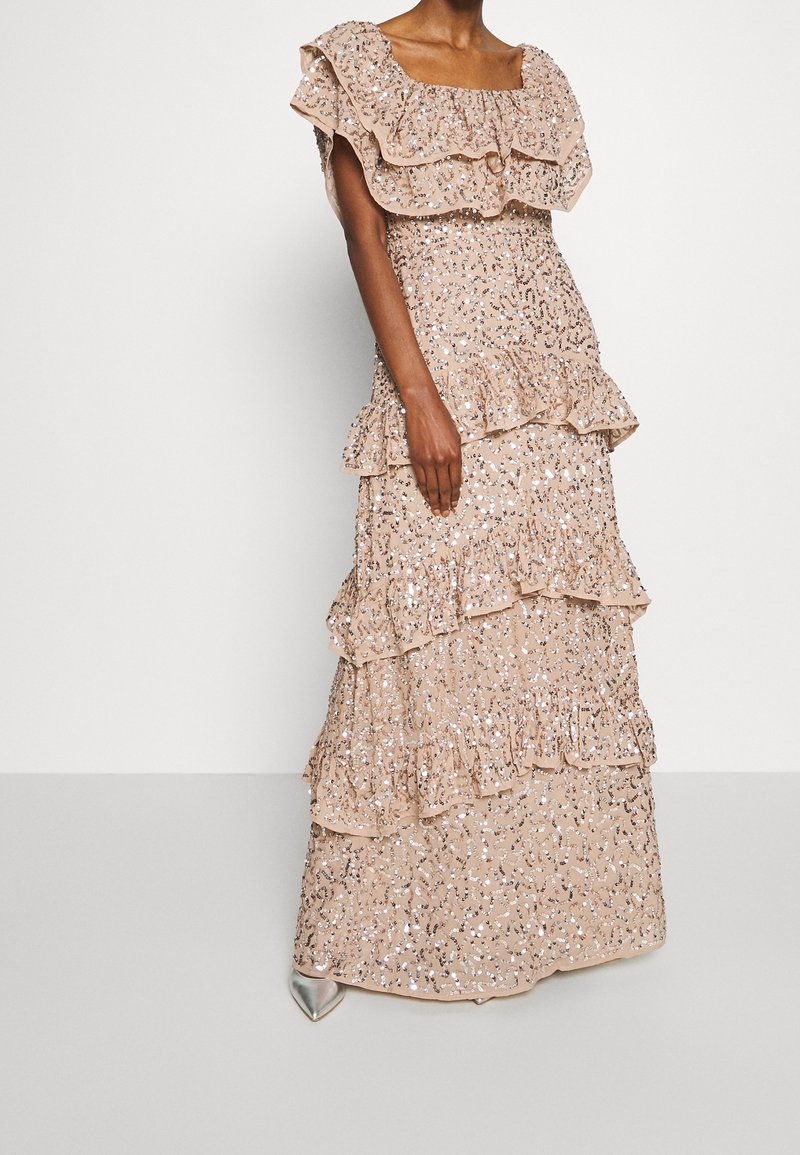 Femme portant une robe beige à volants superposés pailletée, avec des manches épaules dénudées et des talons pointus argentés, sur un fond uni.